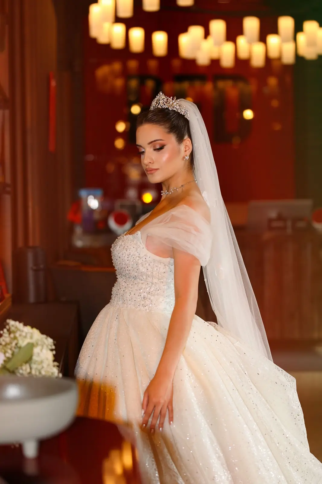 A stunning bride in a sparkling white gown and tiara, posing gracefully indoors.