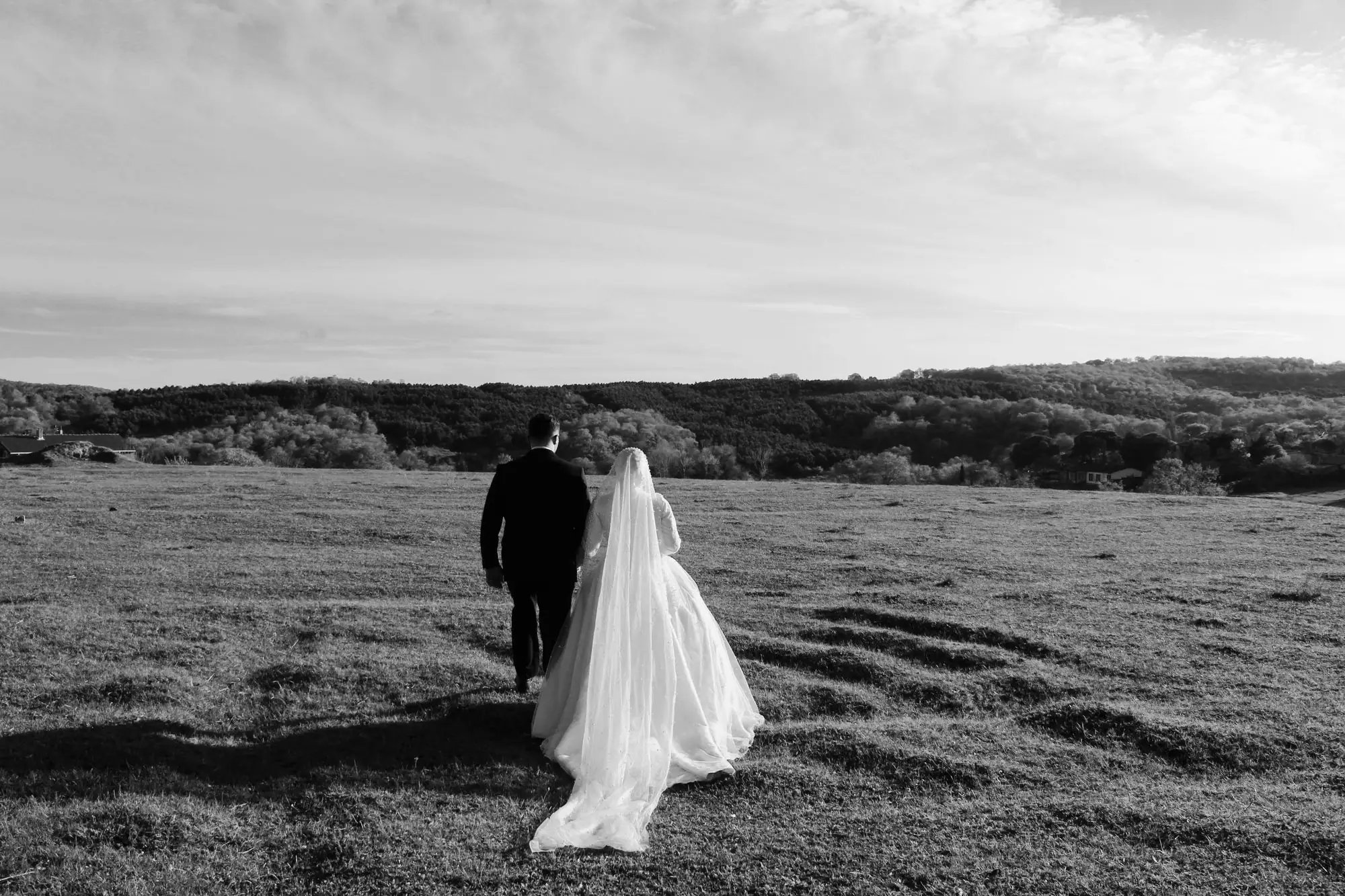 Elegant couple in wedding attire walking in a scenic rural field.