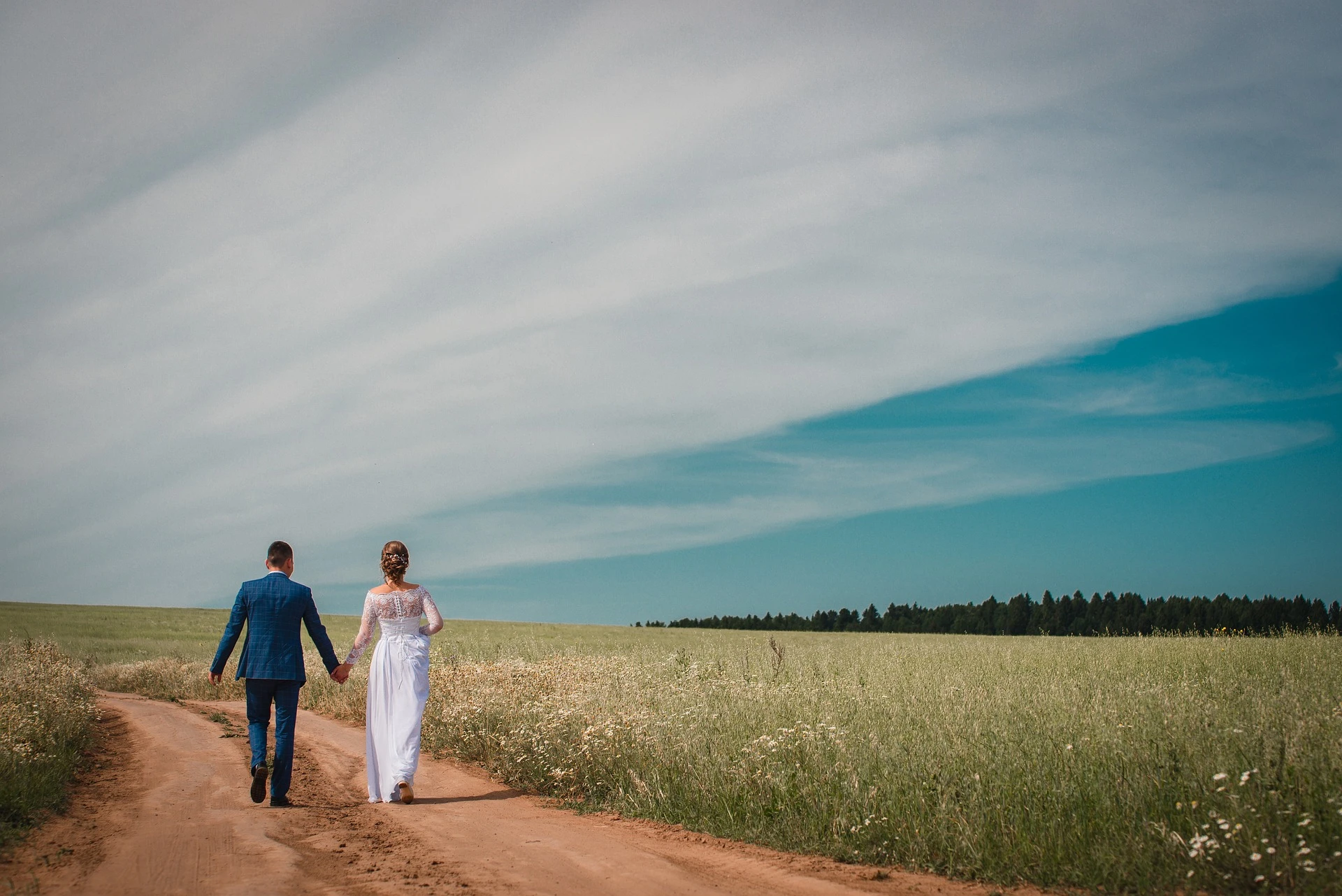 Bride and groom holding hands on a country path beneath a blue sky.