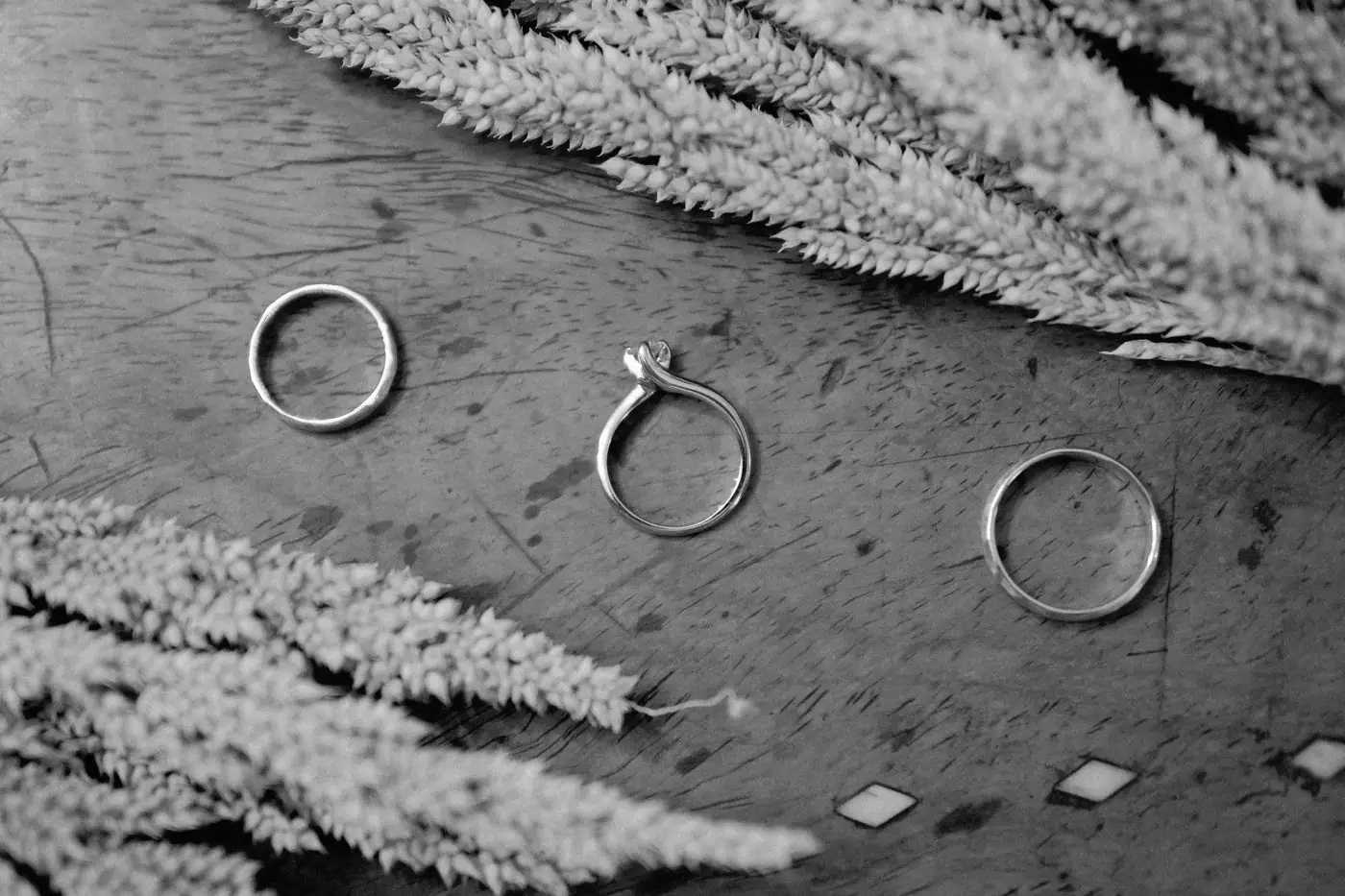 Black and white image featuring wedding and engagement rings on a wooden table with plant accents.