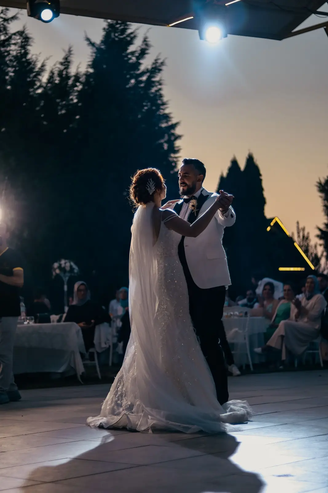 Bride and groom share a romantic first dance outdoors at twilight, surrounded by guests.