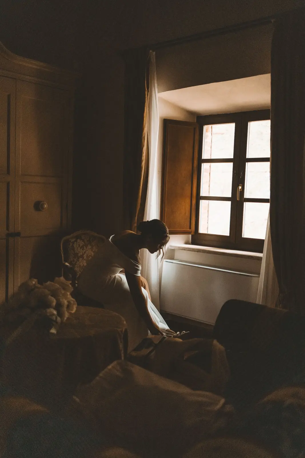 A bride sitting in a softly lit vintage room, adjusting shoes by the window.