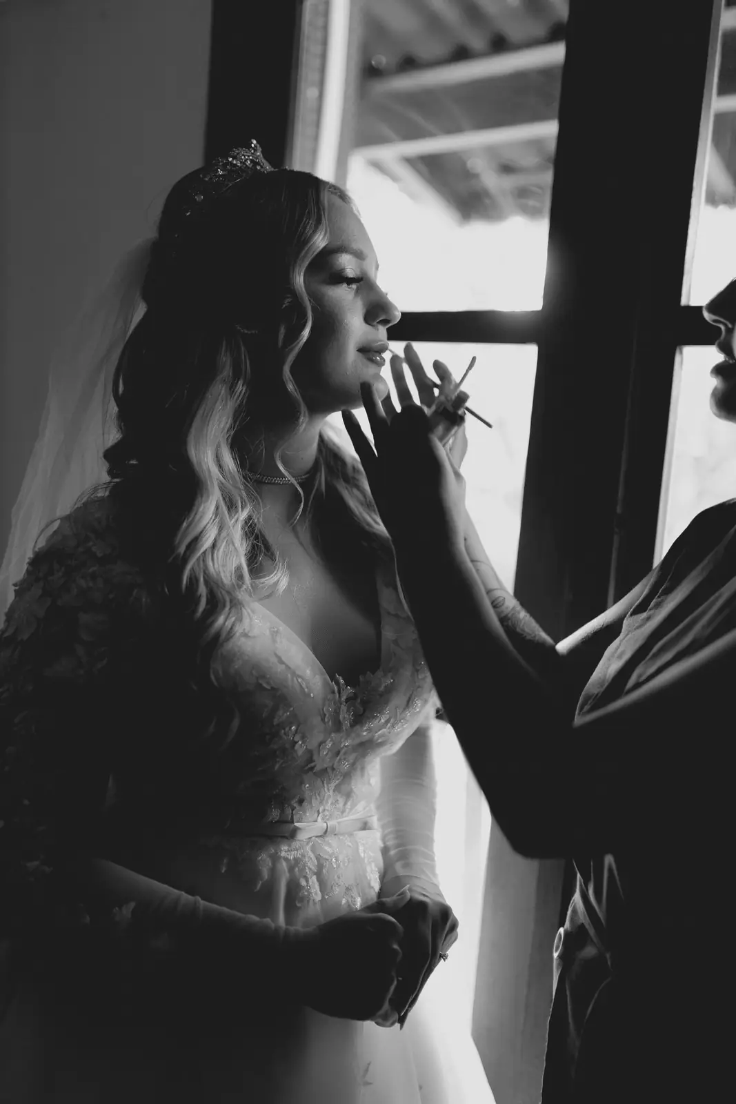 Bride in elegant gown getting makeup done, silhouetted by window light.