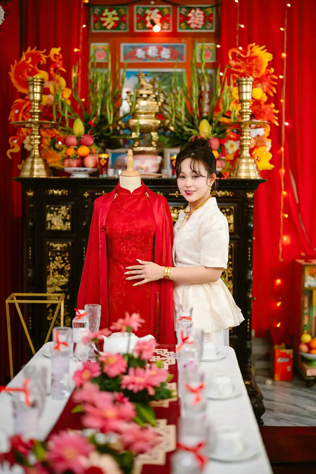 A woman stands beside a traditional Vietnamese wedding dress in a brightly decorated room.