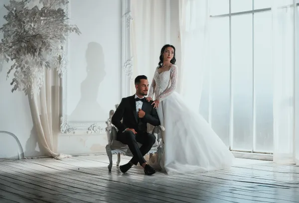 Bride standing beside a seated groom in a bright room with tall windows.