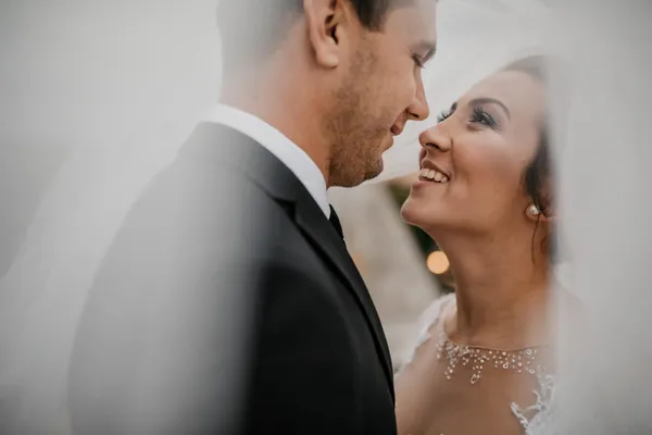 Bride and groom smiling at each other beneath a wedding veil.