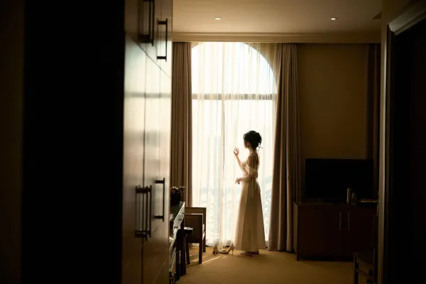 Bride in wedding dress gazing out a sunlit window, creating a serene and reflective mood.