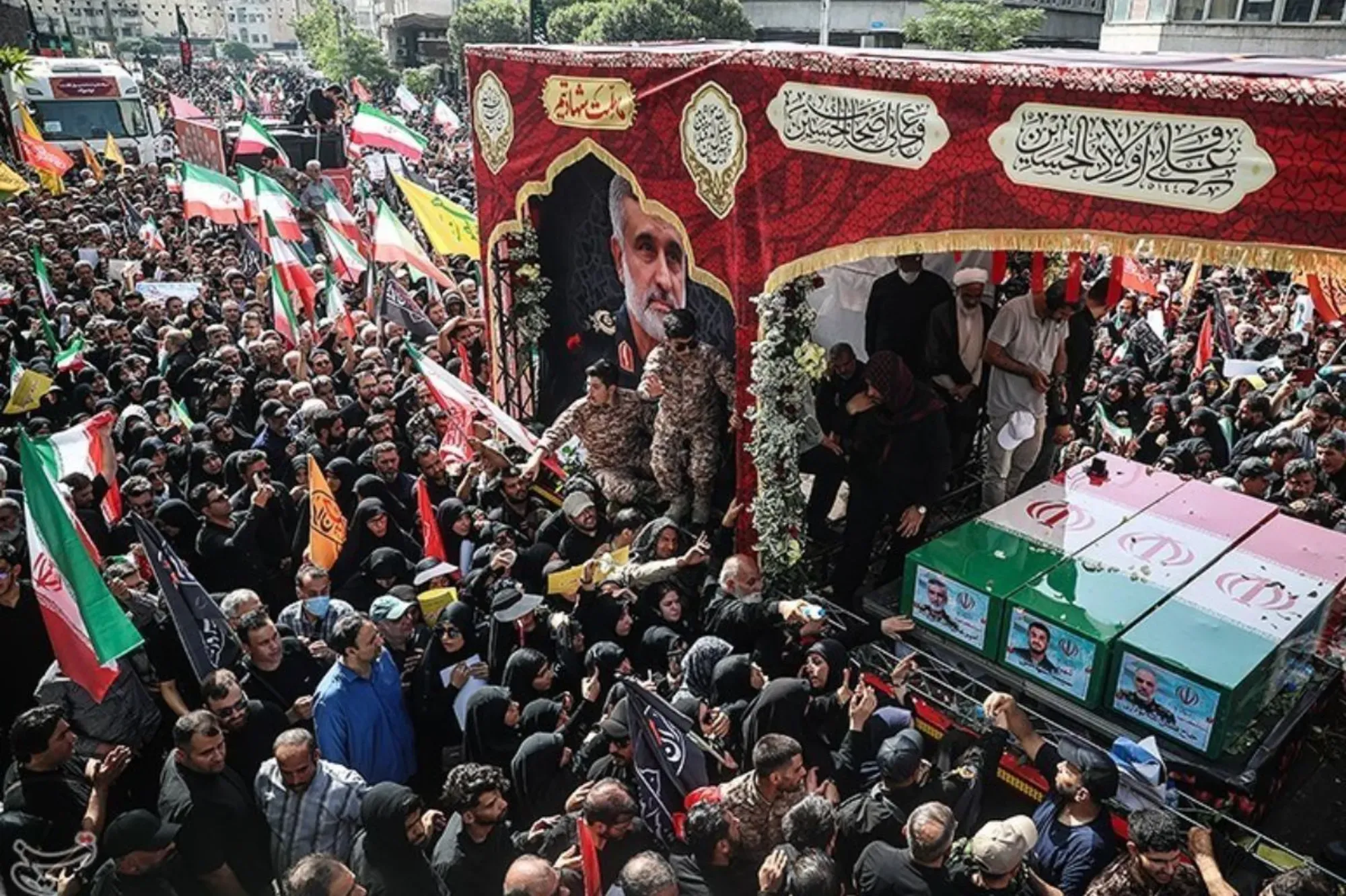 Large funeral procession in Iran with mourners surrounding flag-draped coffins on a vehicle in a crowded city street.