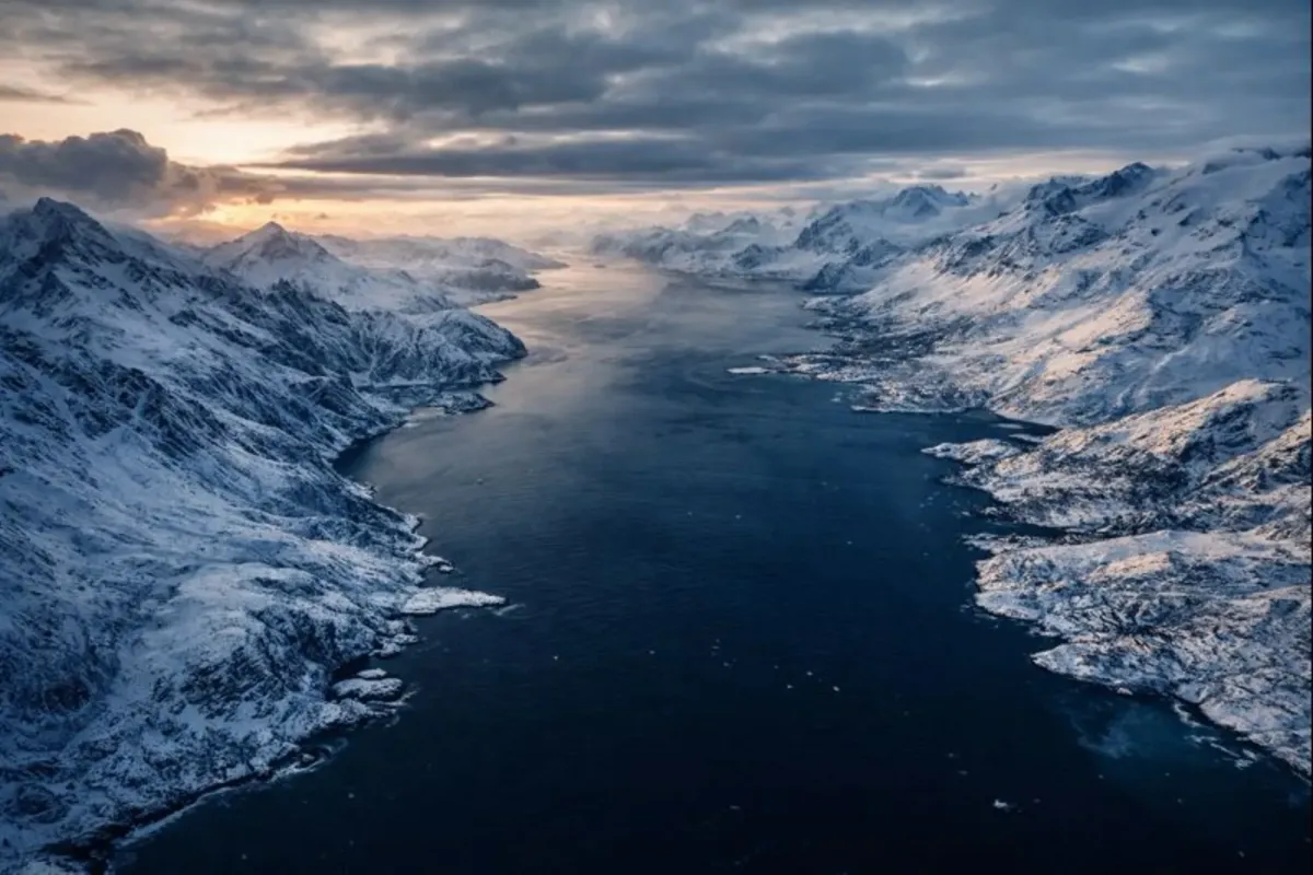 Aerial view of Greenland’s icy coastline with mountains, fjords, and Arctic Ocean under dramatic light.