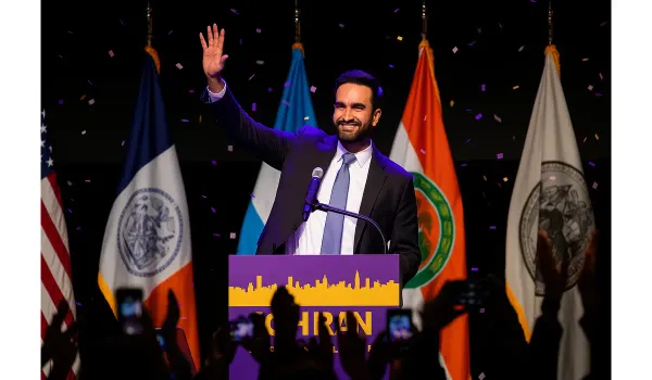 Zohran Mamdani waves to supporters during his victory speech in Brooklyn, surrounded by flags and cheering crowd.