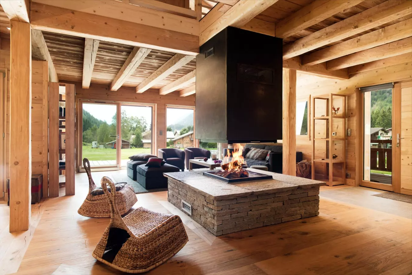 Séjour cosy en bois avec cheminée centrale, grandes baies vitrées et vue dégagée sur la montagne.