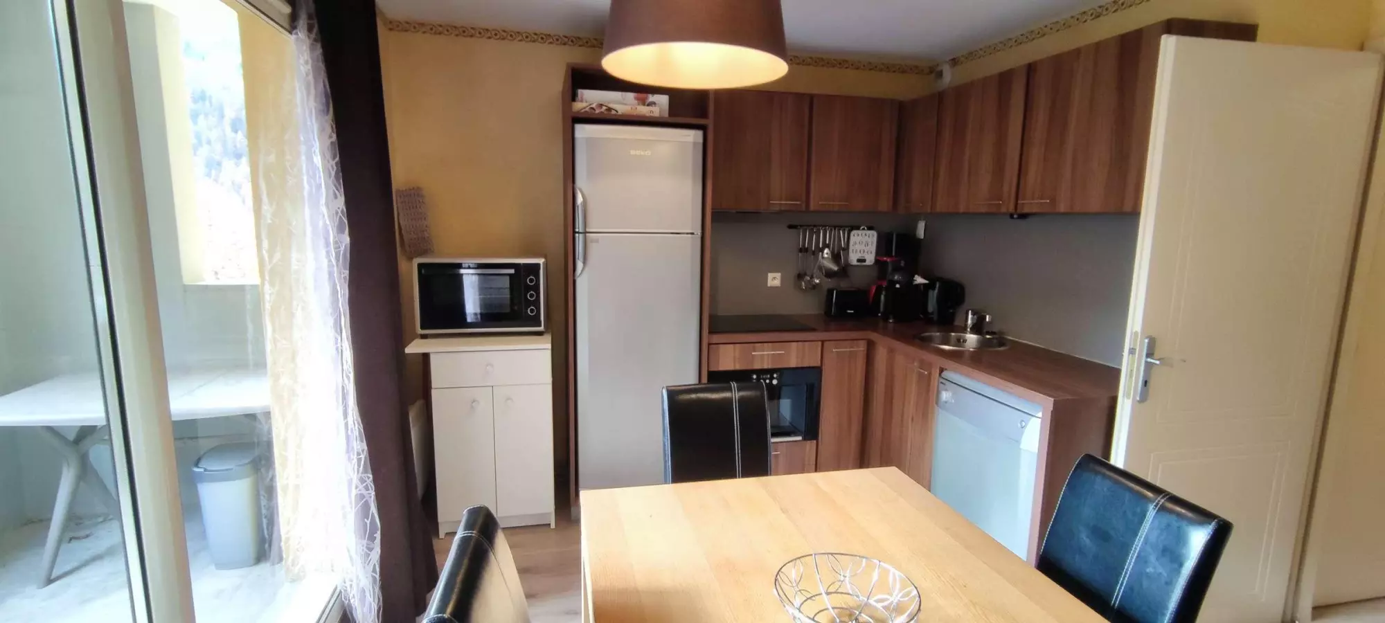 Cuisine équipée avec meubles en bois, électroménagers modernes et coin repas avec table en bois et chaises en cuir noir.