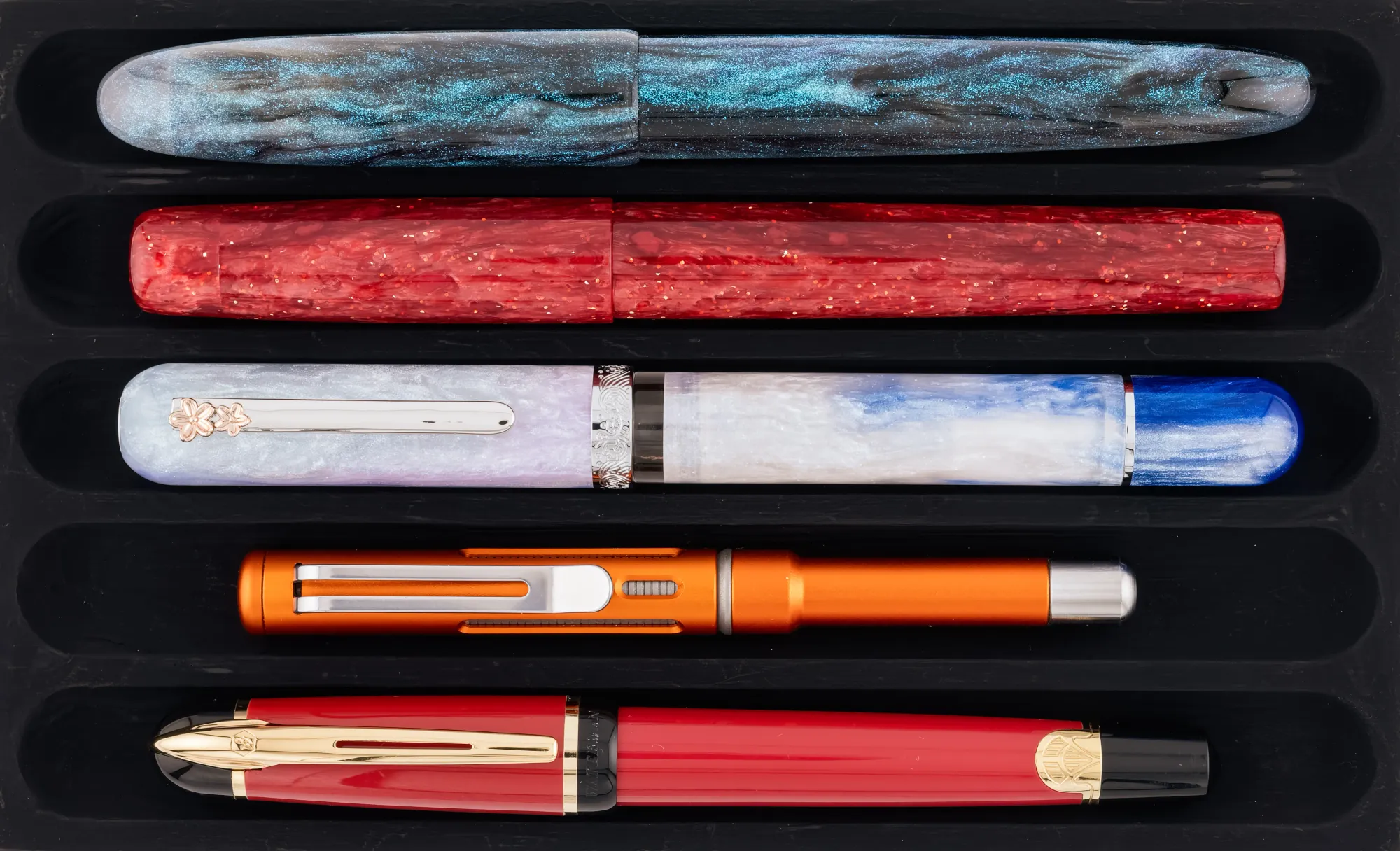 Overhead photo of 5 pens on a matte black pen tray. From top to bottom: Asvine C80, John Garnham JG8, Nahvalur Voyage, Spoke Roady, Waterman Phileas