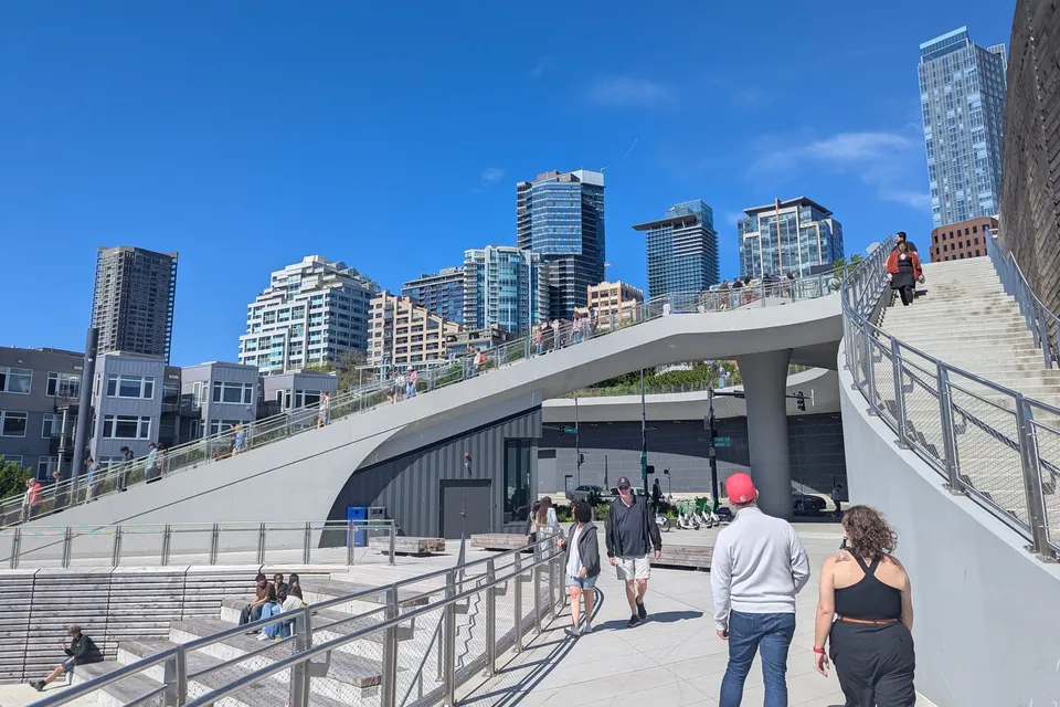 Pedestrians walk through a sunny plaza with Seattle's skyline in the background